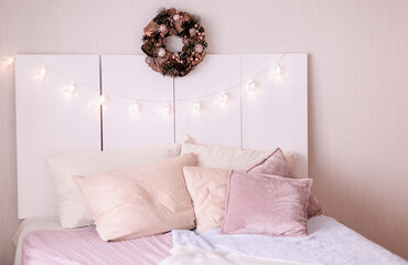 Christmas decoration of the bedroom. The wooden headboard is decorated with a garland of white houses with Scandinavian-style glowing lights and a spruce wreath. Cozy bed with knitted blankets