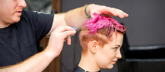 Haircut of dyed short pink wet hair of young caucasian woman by a male hairdresser in a barbershop