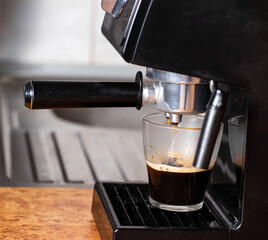 Making coffee in a coffee machine in the kitchen, close-up. Black coffee flowing into a transparent cup on a wooden table top background