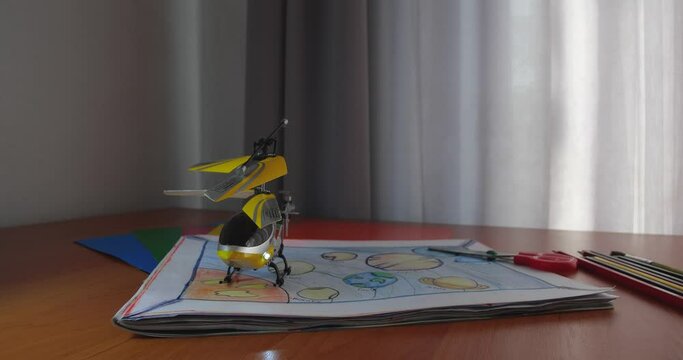 Toy yellow radio-controlled model  helicopter taking off and flying from children's desk with drawings, pencils and scissors making wind. Window is in the background. Slow motion 200 fps