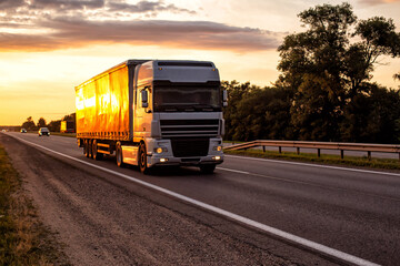 A semitrailer tractor with a tilt semitrailer transports cargo against the backdrop of an evening sunny sunset in summer. The concept of the logistics system and the business of cargo transportation - obrazy, fototapety, plakaty