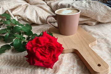 Obraz na płótnie Canvas A large garden red rose is lying on the bed next to a cappuccino mug on a wooden tray. Good festive morning