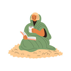 Smiling girl wrapped in a blanket sits on a wool carpet and reads, holding a cup of warm drink