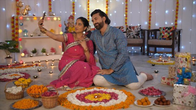 Wide shot of a married couple during Diwali time - Indian festival  decorated house  flower decoration  holy festival. An Indian couple clicking a selfie while sitting on the floor in the living ro...