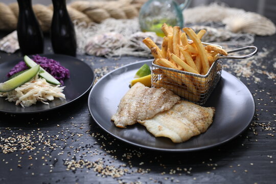 Fried fish fillets served with potato fries in a metal serving basket and salad mix, on black plates, selective focus.
