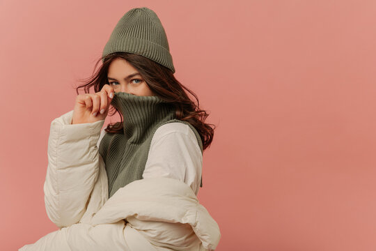 Cheerful young caucasian girl looking into cameras covers her face with collar of sweater on pink background. Brunette with wavy hair wears grey hat, sweater and white jacket. 