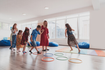 Small nursery school children with female teacher on floor indoors in classroom, doing exercise. Jumping over hula hoop circles track on the floor.