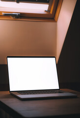Opened modern professional laptop on a wooden table against opened roof window. Concept of working remotely in a cottage. Design mock-up with blank computer screen.