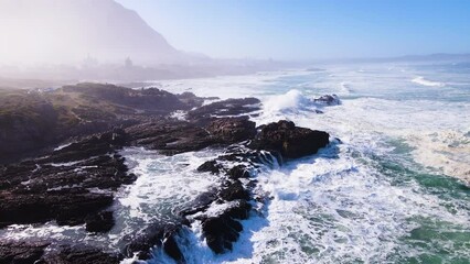 Aerial riser over rocky coastline as waves keep rolling in; Hermanus