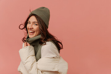 Pretty young caucasian woman laughs looking into camera on pink background with place for text. Brunette with wavy hair wears grey hat, sweater, and white jacket. 