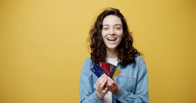 Smiling 20-year-old girl posing isolated on yellow background studio. The concept of people's lifestyle. Looking at the camera, she holds the bank credit card in his hand and shows the winning gesture
