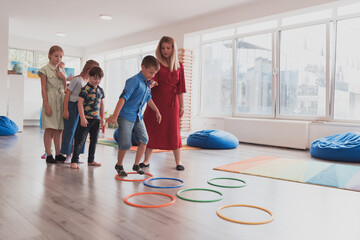 Small nursery school children with female teacher on floor indoors in classroom, doing exercise. Jumping over hula hoop circles track on the floor.