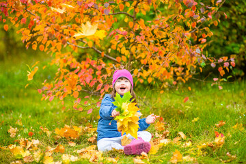 Happy young girl with Downs syndrome sits on autumn leaves at park and looks up on leaf fall
