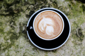Cappuccino coffee with a pattern of milk foam in a black cup on a stone background with moss.