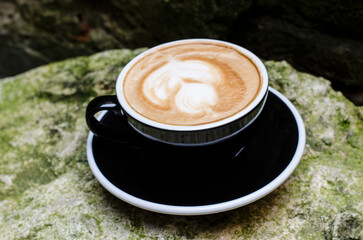 Cappuccino coffee with a pattern of milk foam in a black cup on a stone background with moss.