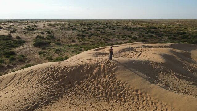 drone is circling around a girl in a long leopard dress stands on a high dune in a desert with grass in summer in Astrakhan