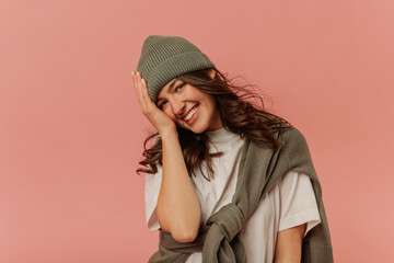 Pretty caucasian young woman smiles looking into camera holding head on pink background. Brunette with wavy hair wears grey hat, sweatshirt, and white T-shirt. Lifestyle, emotions, leisure concept. 