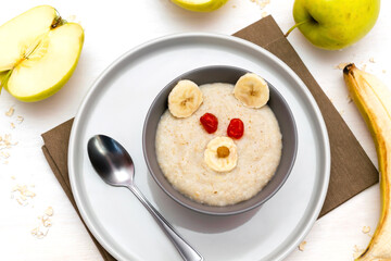 Funny cute kids childrens baby's healthy breakfast lunch oatmeal porridge in bowl look like bear face decorated with apple, banana, dried berry fruits. dessert food art on white wooden table