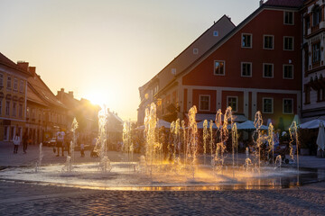 Flowing fountain in center of Maribor in golden hour. Splashing water on square in old city in Slovenia in sunset light. Historical building in turistic town. - Powered by Adobe