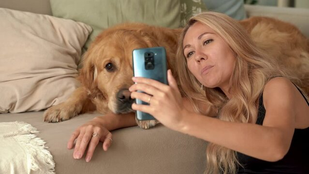 Caucasian blonde woman sitting on carpet with dog Labrador. Happy young student pet owner doing selfie picture, talking and laughing with beautiful Labrador dog at home. 4k footage