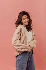 Stylish young brunette caucasian woman is smiling looking at camera on pink background. Hottie wears white shirt, beige jacket and jeans. Lifestyle concept 