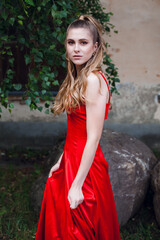 A cute young girl with blond flowing long hair and beautiful make-up, in a long red dress, stands on the street of the old city. There is a tree nearby.