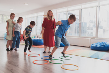Fototapeta premium Small nursery school children with female teacher on floor indoors in classroom, doing exercise. Jumping over hula hoop circles track on the floor.