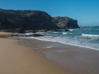 View of rocks and cliff at Praia das Furnas golden sand beach with turquoise water of Atlantic ocean near Vila Nova de Milfontes, Vicentine Coast Natural Park Portugal. Sunny day, blue sky