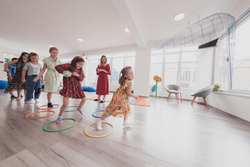 Small nursery school children with female teacher on floor indoors in classroom, doing exercise. Jumping over hula hoop circles track on the floor.