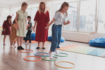 Small nursery school children with female teacher on floor indoors in classroom, doing exercise. Jumping over hula hoop circles track on the floor.