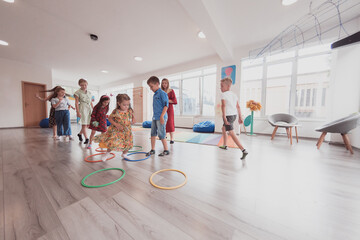 Obraz premium Small nursery school children with female teacher on floor indoors in classroom, doing exercise. Jumping over hula hoop circles track on the floor.