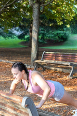 View of a beautiful girl doing push-ups on a park bench. Sporty girl doing push-ups on a bench in a nice park
