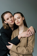 young and pretty lesbian women in black and beige blazers looking at camera isolated on grey.