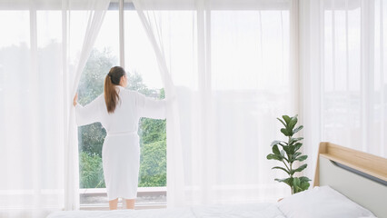 Woman wearing bathrobe opens curtains in bedroom and see around the outdoors. Welcome new sunny day, happy positive young woman wakes up and looks in the window at home or hotel feeling optimistic.