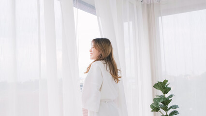 Woman wearing bathrobe opens curtains in bedroom and see around the outdoors. Welcome new sunny day, happy positive young woman wakes up and looks in the window at home or hotel feeling optimistic.
