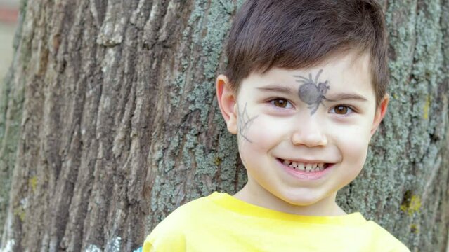 portrait of cute smiling little boy on tree trunk wooden texture.kid with spider and black spider web drawing on forehead and cheek.halloween trick or treat holiday concept.face painting safe washable
