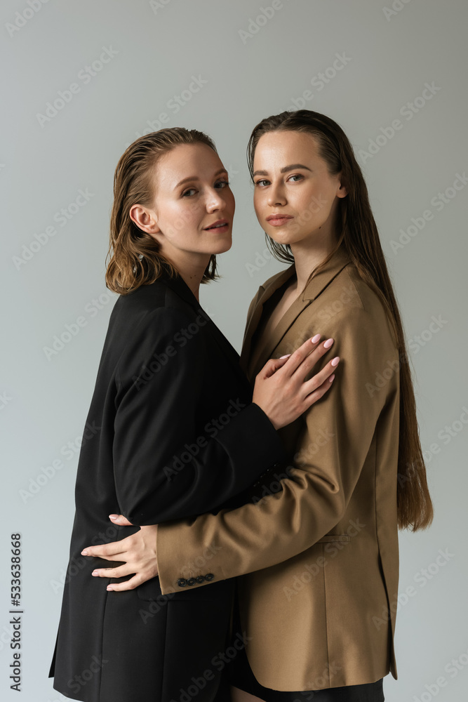 Wall mural sensual lesbian couple in black and beige blazers embracing and looking at camera isolated on grey.