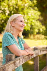 Portrait Of Casually Dressed Mature Or Senior Woman Leaning On Fence On Walk In Countryside