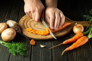 The chef cuts raw carrots with a knife on a wooden cutting board to prepare a vegetarian dish. Peasant food