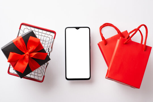 Cyber monday concept. Top view photo of smartphone red paper bags and giftbox with ribbon bow in shopping cart on isolated white background with copyspace