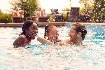 Smiling Mixed Race Family On Summer Holiday Having Fun Splashing In Outdoor Swimming Pool