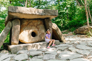 Little girl sit near ancient dolmen in green park on sunny day. Tourists look at natural sights on summer vacation. Single chamber megalithic tomb, old stone structure. Tourism, beautiful place.