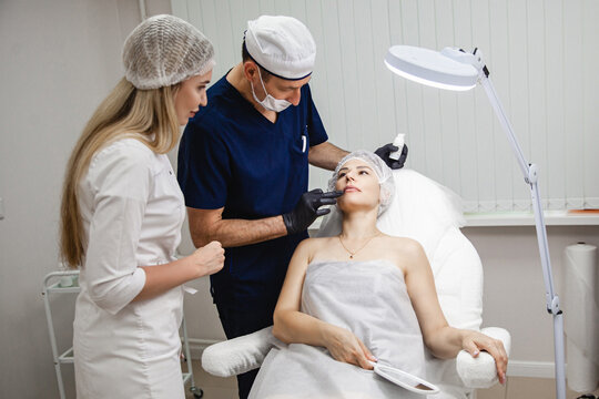 A man doctor with a girl assistant examine the patient's woman before a lip augmentation procedure in a cosmetology clinic