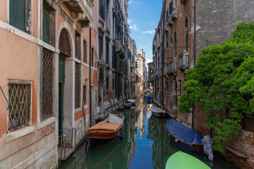 Fototapeta na wymiar Boats near vintage brick walls of houses on the water surface of a narrow canal street in Venice, reflections and sky on a Venetian street, a tree on the bank of a Venice canal on a sunny day