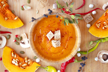 Warm and healthy pumpkin soup with croutons in wooden plate surrounded by garlic, red pepper, mushrooms, pumpkins, berries on wooden table. Top view. Flat lay. High quality photo