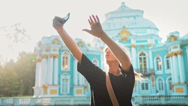 Beautiful caucasian teenage girl walks in city center and takes selfie against of cathedral or architectural building. Female poses, touches hair and happy smiling. Photos with attractions in summer.