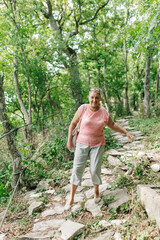 Naklejka na ściany i meble Middle aged woman singly go down stone path without looking at feet in green forest. Tourist with interest walk along dangerous route. Hiking in mountains, active recreation, vacation.