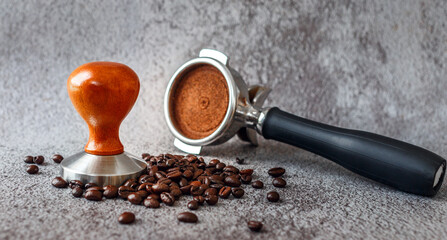 Equipment in a coffee shop of barista coffee tool portafilter with tamper and dark roasted coffee beans on gray background