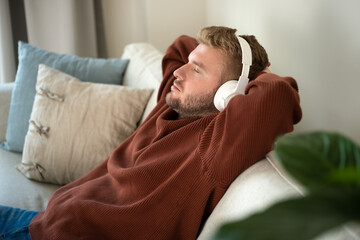 Young caucasian blond curly hair man listening to music with big white handsfree headphones sitting...