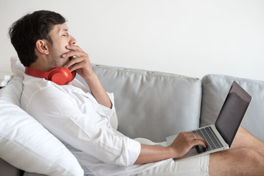 Young Asian Man Relaxing On Comfortable Sofa In Living Room With Laptop Computer At Home Bored Yawning Tired.